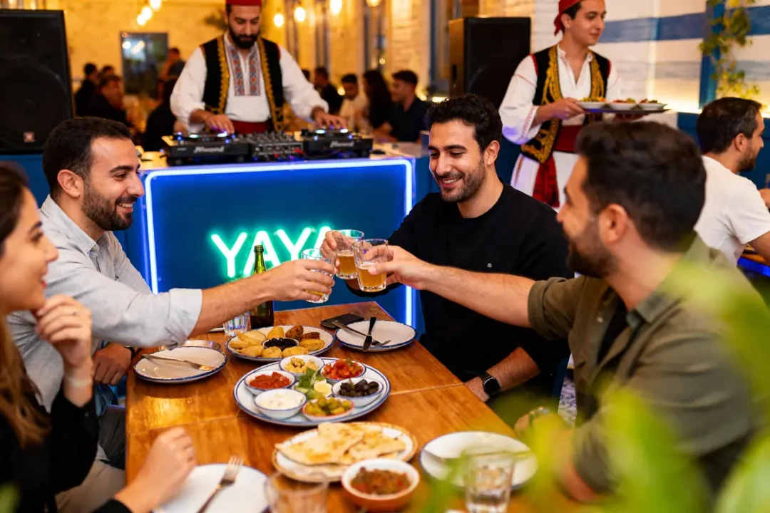 Des amis trinquent autour d'un repas convivial dans un restaurant animé, avec DJ et serveur en tenue traditionnelle.