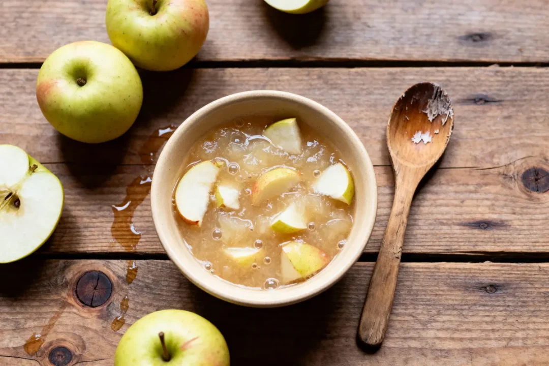 Bol de compote de pommes avec morceaux, cuillère en bois, pommes fraîches sur une table en bois rustique.