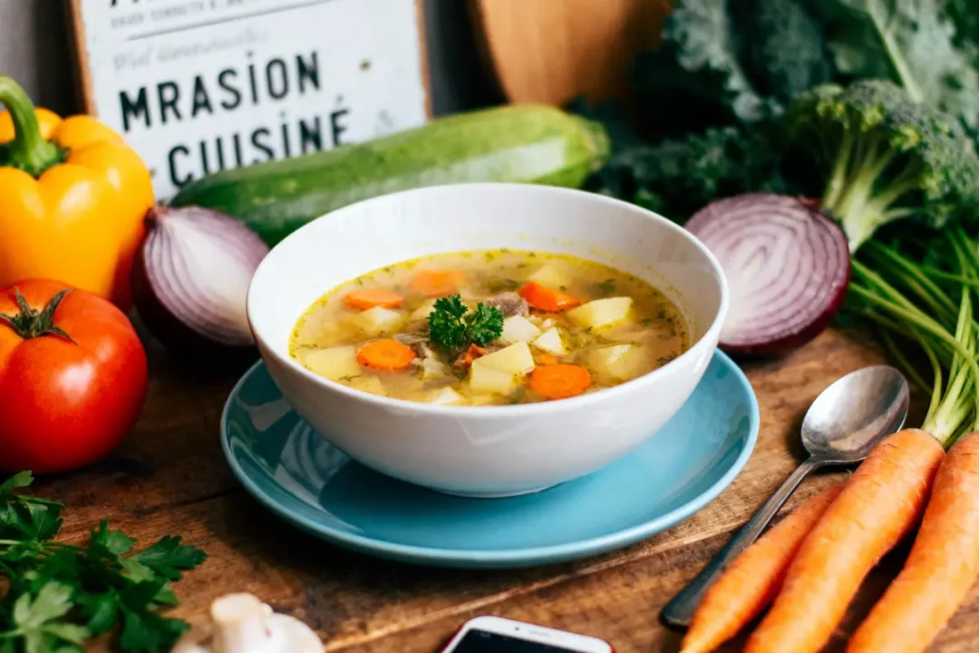 Bol de soupe aux légumes avec carottes, pommes de terre, entouré de légumes frais sur table en bois.