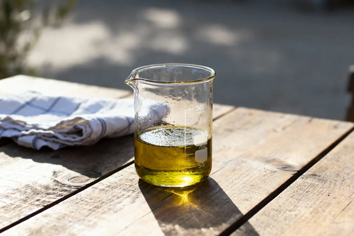 Bécher contenant du liquide jaune sur table en bois, à côté d'un torchon blanc, extérieur ensoleillé.