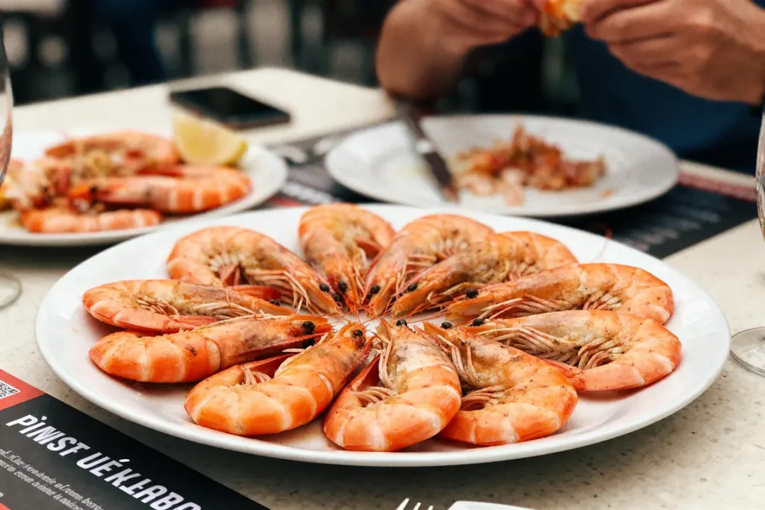 Assiette de crevettes cuites, disposées en cercle, sur table avec citron et personne mangeant en arrière-plan.