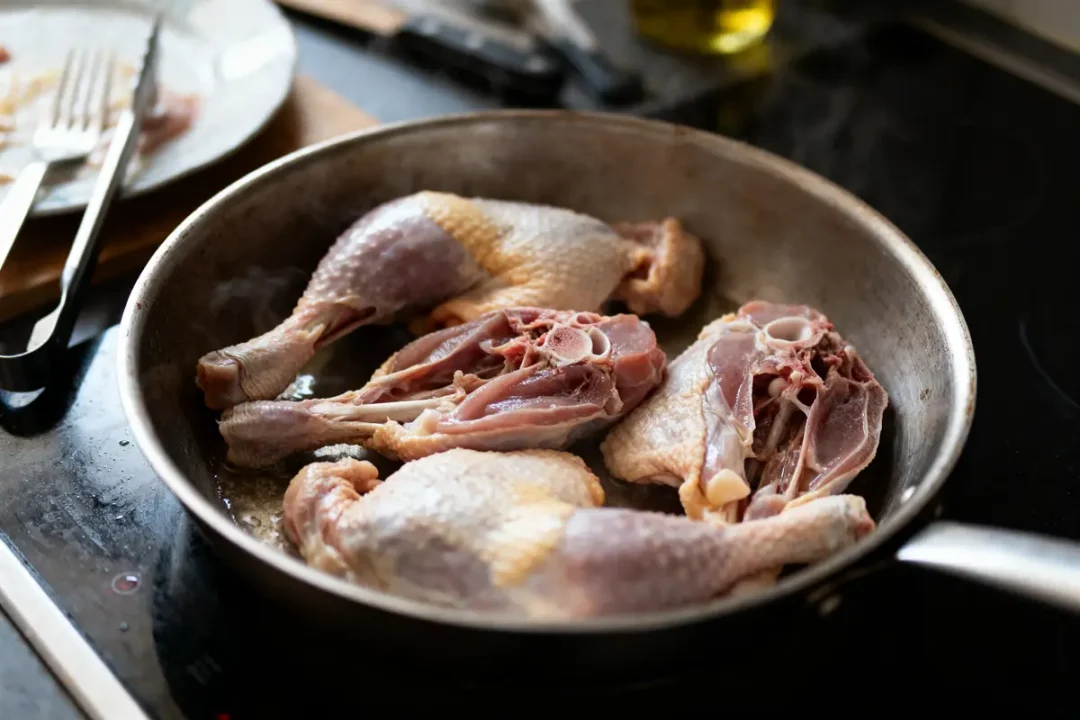 Cuisses de poulet crues dans une poêle en métal, sur une cuisinière. Deux côtelettes visibles en arrière-plan.