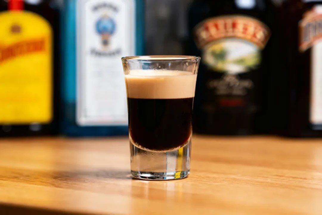 Verre à shot avec cocktail en strates foncées et crémeuses, devant des bouteilles floues sur le fond.