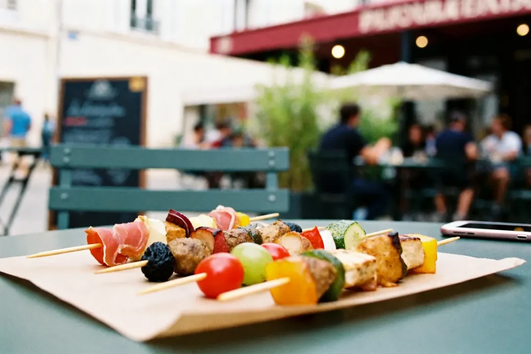 Brochettes colorées sur table extérieure, arrière-plan flou avec personnes assises, enseigne restaurant en arrière-plan.
