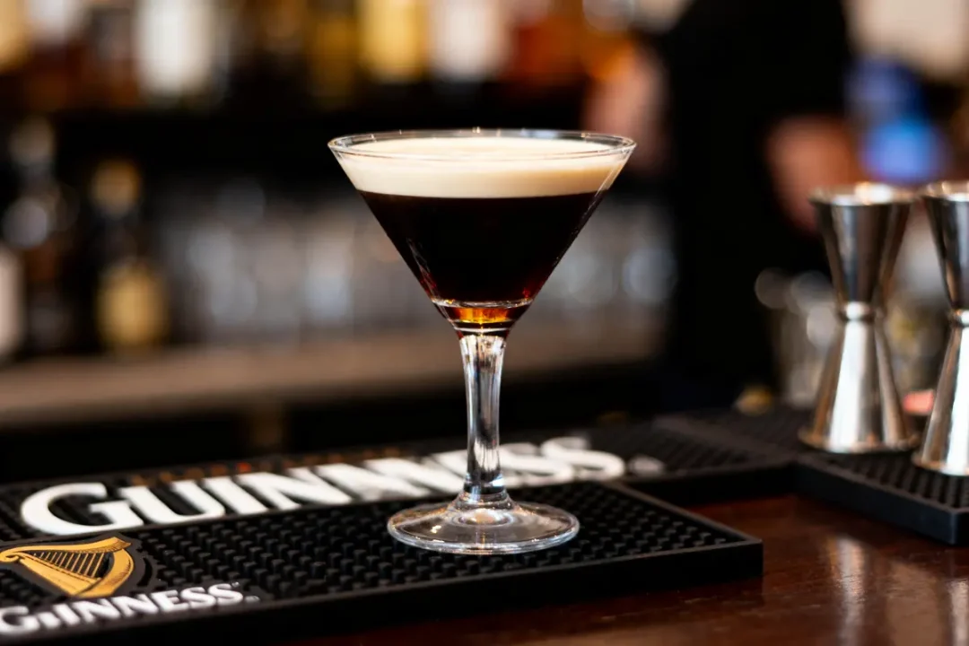 Cocktail espresso martini sur un bar, avec mousse blanche et arrière-plan flou de bouteilles et accessoires.