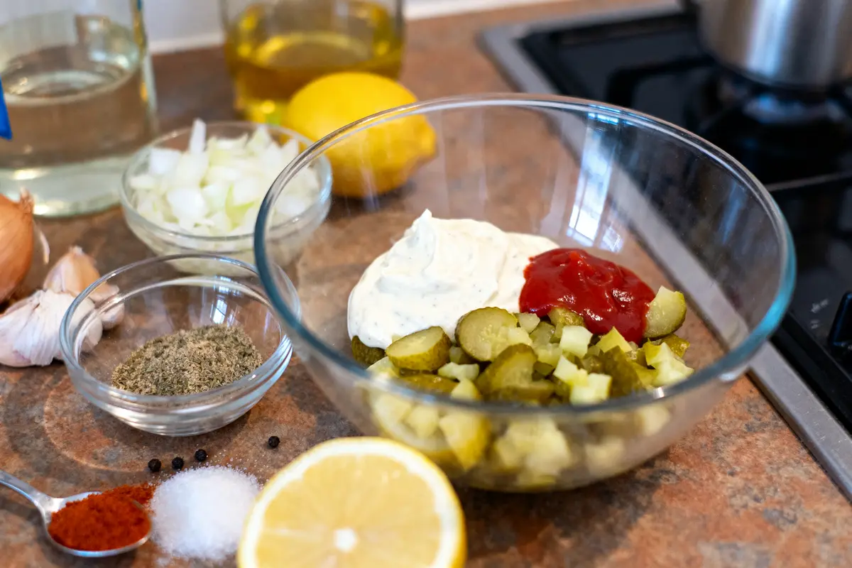 Bol de préparation avec cornichons, mayonnaise, ketchup, épices, oignon et citron sur un plan de travail.