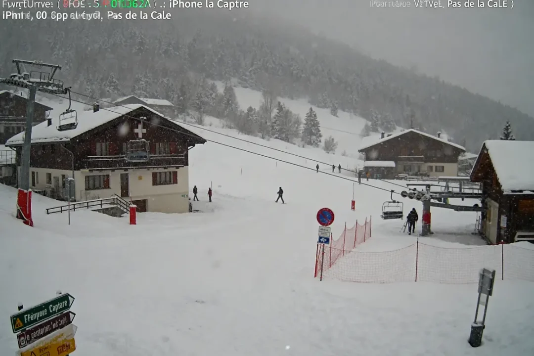 Chalets enneigés, téléski, skieurs, panneau indicateur, forêt en arrière-plan sous la neige.