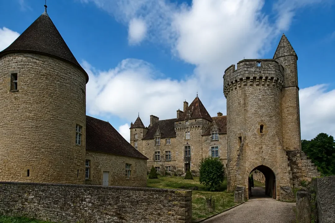 Château médiéval en pierre sous un ciel bleu avec nuages, créneaux et tour ronde.