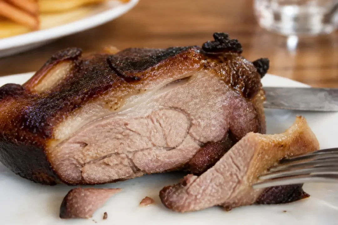 Tranche de viande de canard rôti, peau croustillante, sur assiette blanche avec fourchette et couteau.