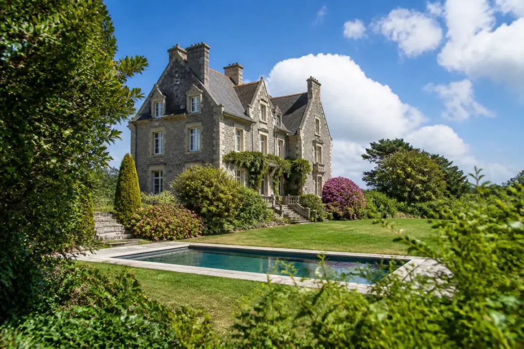 Manoir en pierre avec piscine, entouré de verdure, ciel bleu et nuages blancs en arrière-plan.