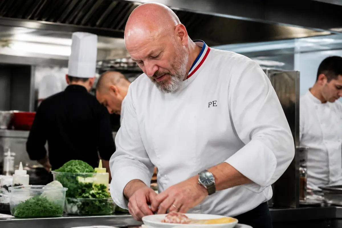 Chef cuisinier préparant un plat dans une cuisine professionnelle, entouré de chefs en tenue blanche.
