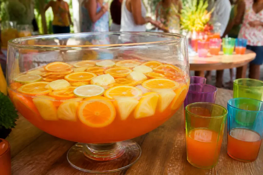 Grande coupe de cocktail aux fruits, avec tranches d’agrumes, entourée de verres colorés. Scène de fête conviviale.