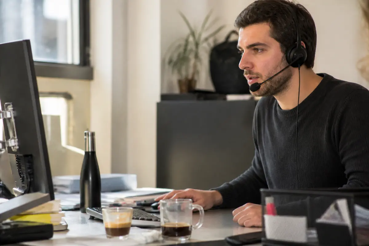 Un homme en casque devant un ordinateur, buvant un café. Cadre de bureau moderne.